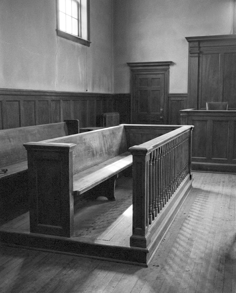 An empty wooden dock with a worn rail inside a panelled courtroom, a single shaft of light falling across it.